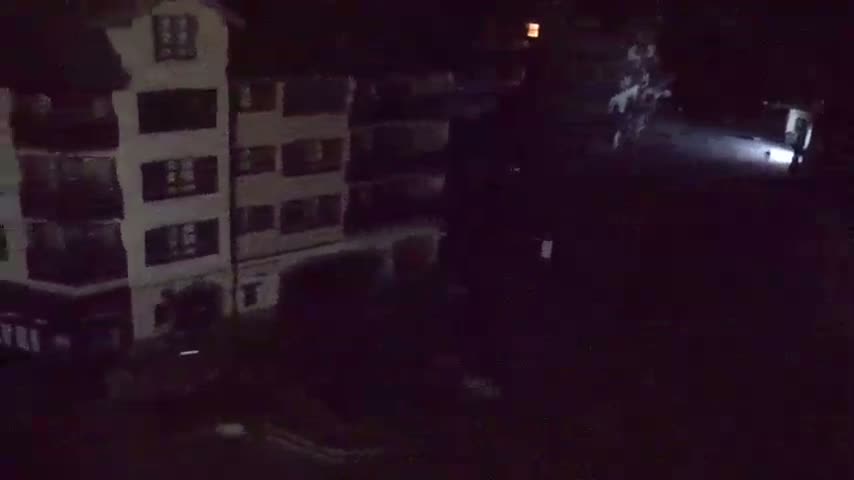 A nighttime, low-light view shows a multi-story building and a dark, possibly empty, area in front of it.