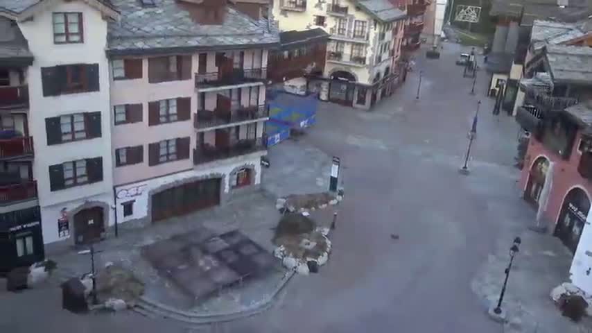 An empty town square, surrounded by multi-story buildings in a muted color palette, on an overcast day.