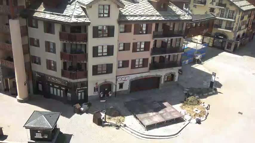 A paved plaza with several buildings, a small stage, and piles of debris is viewed from above on a sunny day.