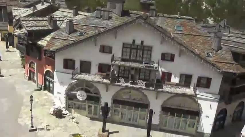 An aerial view shows a cluster of buildings with slate roofs and arched windows, situated in a sunlit area.