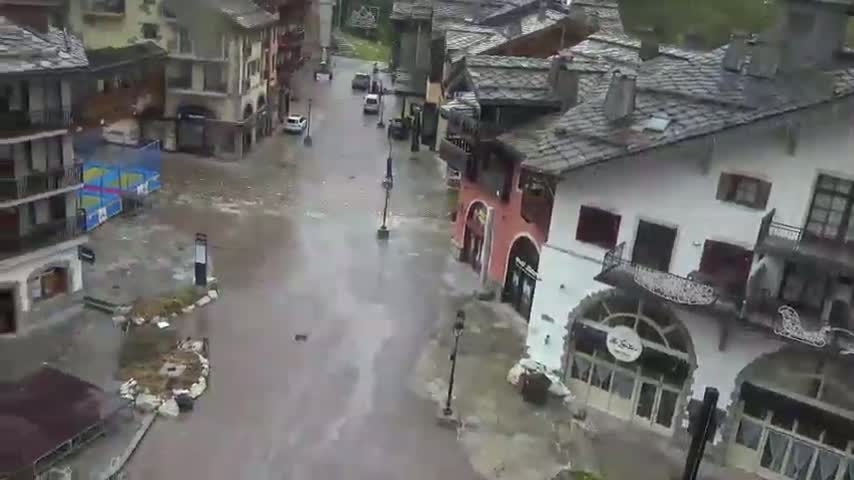 A rain-soaked street in a village with stone buildings is partially flooded, with a few cars visible.