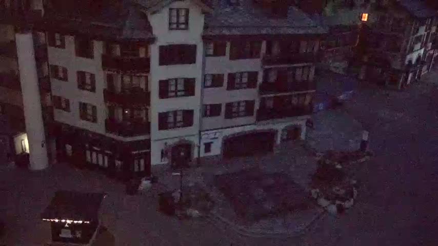 An aerial nighttime view shows a town square with a multi-story building and several smaller structures surrounding it, under dark conditions.