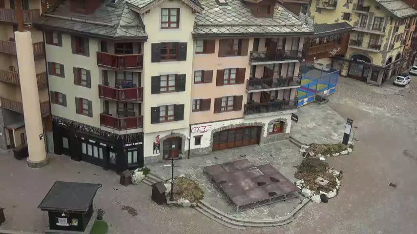 A mostly empty town square with several multi-story buildings, some with balconies, and a dark-colored structure resembling a stage or platform in the center, all under overcast skies.