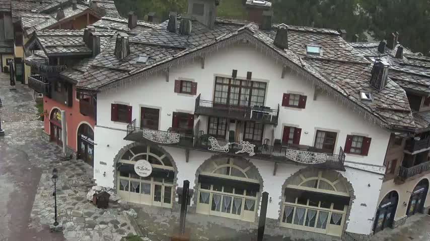 A multi-story building with a slate roof and arched windows sits in a village, and icicles hang from its eaves in overcast weather.