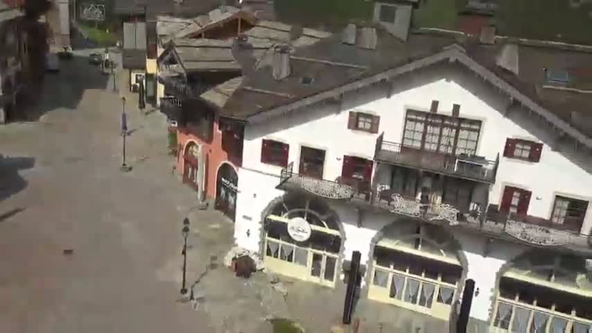 An aerial view shows a plaza and several buildings with balconies in a village on a sunny day.