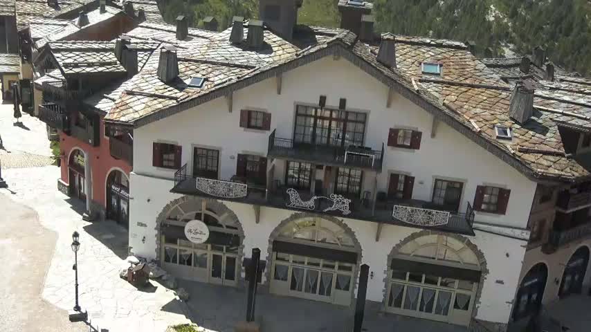 A large, white building with a sloped, slate roof and arched windows, situated among smaller buildings in a sunny, mountainous area.