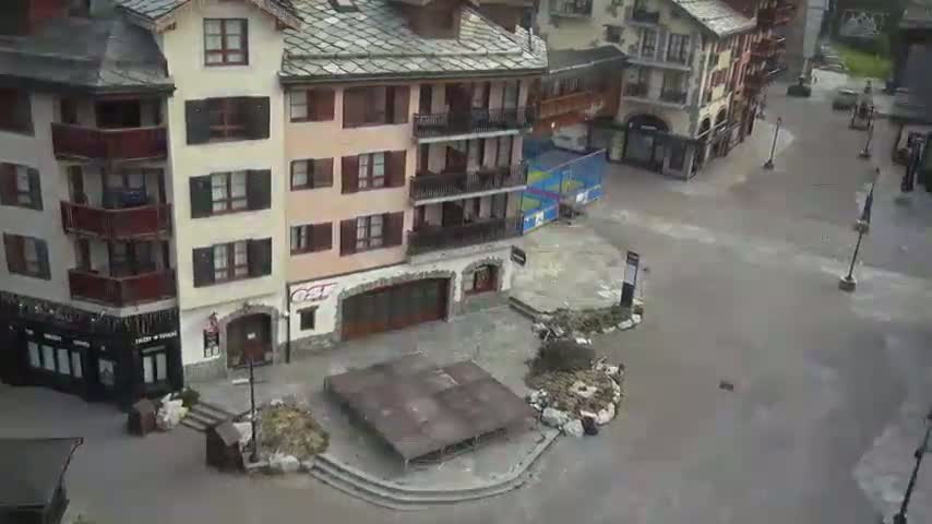 A mostly empty town square with several buildings, some with balconies, is partially flooded under an overcast sky.