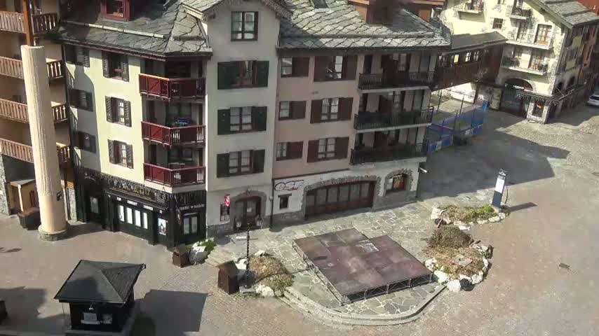 A paved plaza, under sunny conditions, is surrounded by several multi-story buildings with balconies and dark roofs.