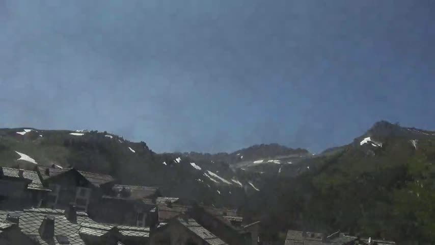 A hazy, sunny day reveals a village of dark-roofed houses nestled at the base of snow-dusted mountains.