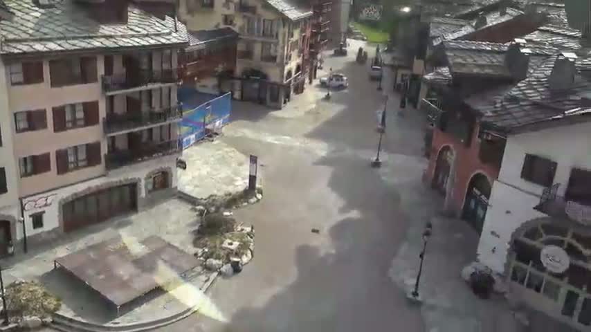 A mostly empty village square with several multi-story buildings is flooded with muddy water.