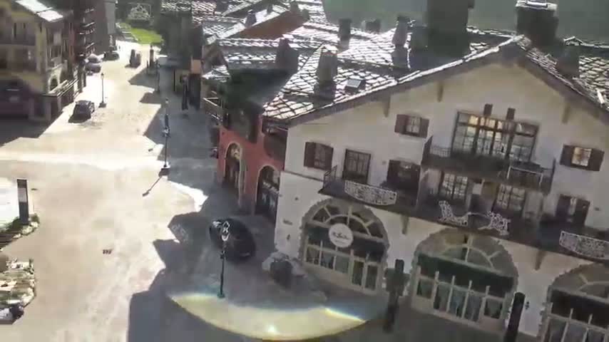 An aerial view shows a village square with several cars and buildings under a sunny sky.