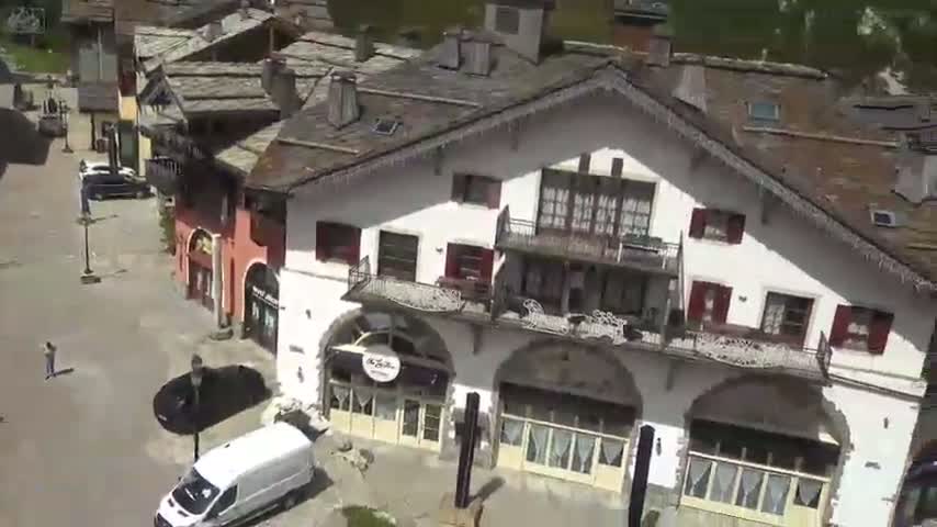 An aerial view shows a village street with several multi-story buildings, a white van, and a few cars parked along the street, under sunny conditions.