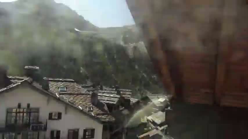 A misty, sunny day reveals a village nestled in a mountain valley, with water spraying from an unseen source.