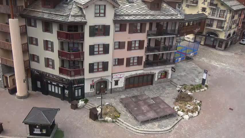 An aerial view shows a paved plaza in a village of multi-story buildings with balconies, a small stage, and landscaping; the weather appears overcast.