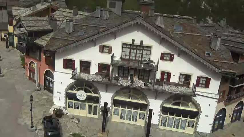 An aerial view shows a cluster of buildings with dark slate roofs and icicles hanging from the eaves, situated in a village on a sunny day.