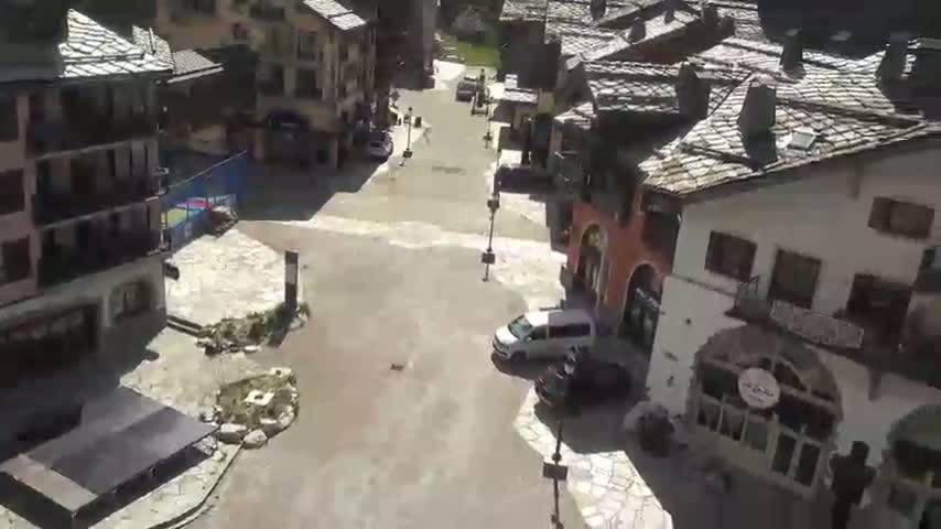 A mostly sunny day reveals a village square flooded with shallow water, where a few vehicles are parked amongst stone buildings with slate roofs.