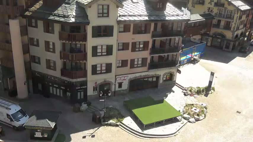 A sunny view of a plaza with a grassy stage and several multi-story buildings.