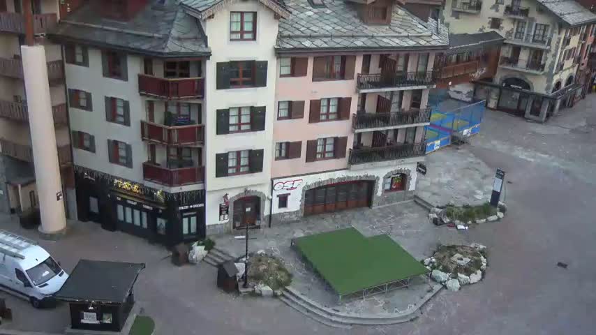 An aerial view shows a mostly empty plaza in a village of multi-story buildings, with a small covered area and a green artificial turf patch visible in the center.