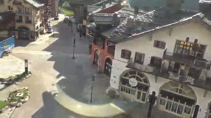 A mostly empty village square with several buildings, some with balconies, is partially flooded under a sunny sky.