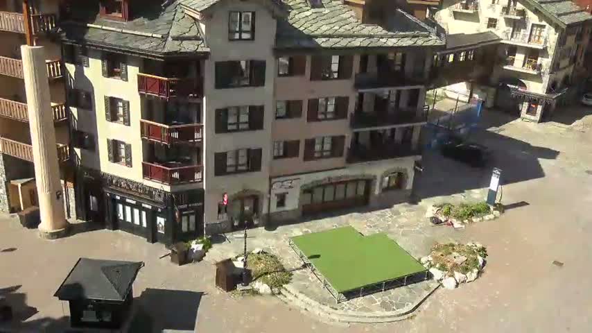 A sunlit plaza with a green temporary stage sits between several multi-story buildings with balconies.