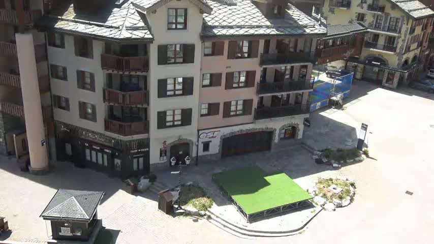 An aerial view shows a sunny plaza with several multi-story buildings, one of which has a green artificial turf area in front of it.