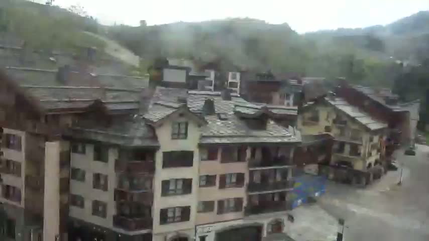 A cluster of multi-story buildings with slate roofs sits in a mountainous area on an overcast day.