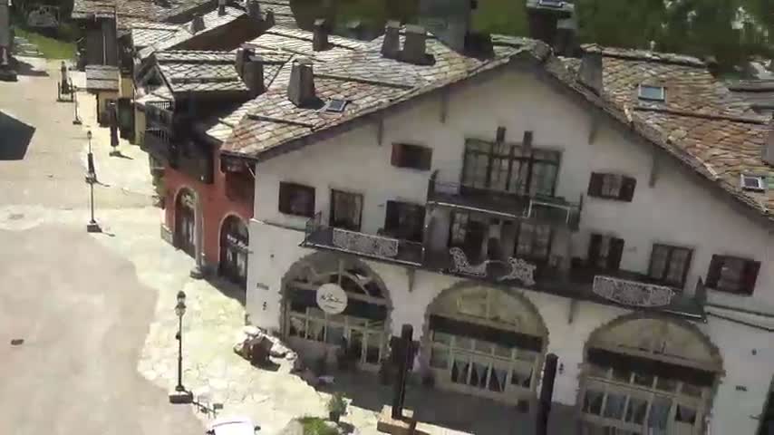 An aerial view shows a village of white buildings with dark slate roofs under a sunny sky.