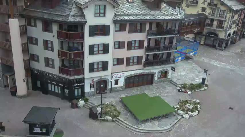An aerial view shows a town square with a small artificial turf area, surrounded by several multi-story buildings in a mountainous area on a cloudy day.