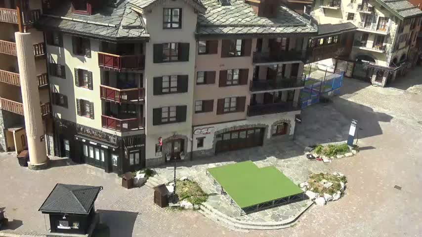 An aerial view shows a paved plaza with a small, raised green stage, surrounded by several multi-story buildings in a sunny, outdoor setting.