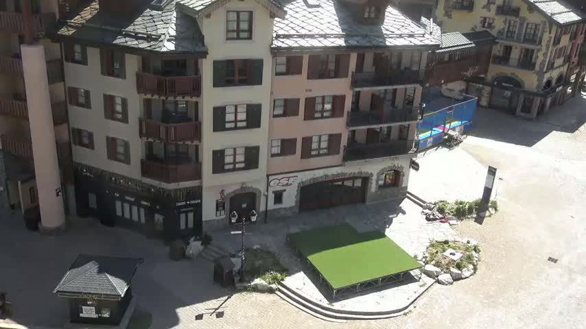 A sunny outdoor plaza with a multi-story building, a small kiosk, and a raised green platform.