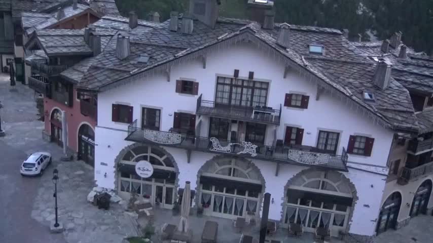 An aerial view shows a white building with dark slate roofs and icicles, situated among other buildings in a village on an overcast day.