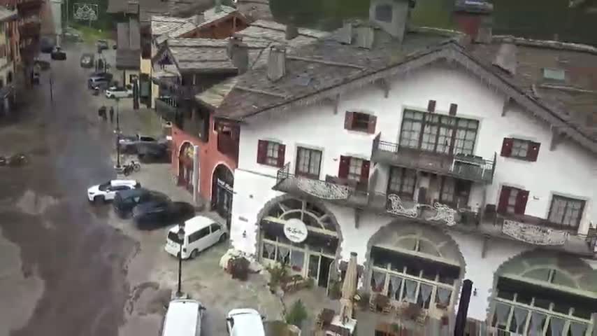 A flood of muddy water covers a street in a village of European-style buildings, with several cars parked along the sides.