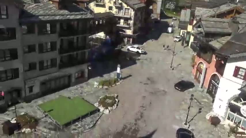 A mostly empty town square, partially flooded, is surrounded by European-style buildings under a sunny sky.