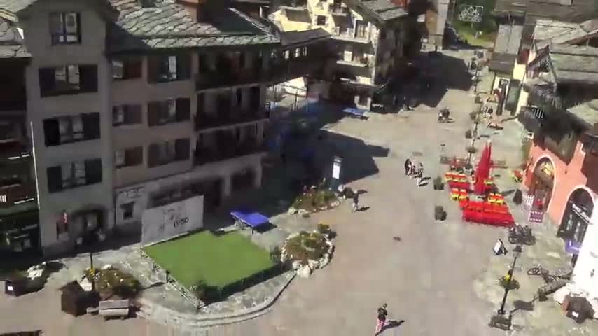 An aerial view of a village square with several buildings, a small green area with a ping pong table, and a few people scattered throughout on a sunny day.
