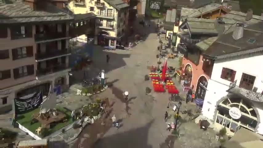 An aerial view shows a village square with several multi-story buildings, some people walking around, and outdoor seating areas under a sunny sky.