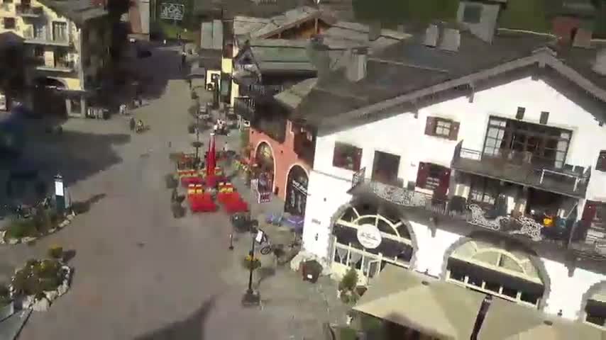 A sunny day reveals a village square with several buildings, outdoor seating, and a few people walking and cycling.