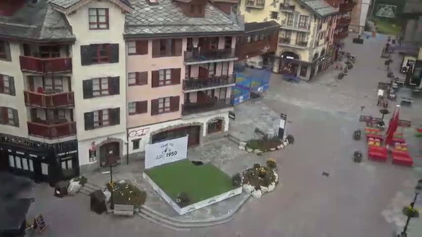 A sunny day reveals a village square with several buildings, outdoor seating, and a few people walking and cycling.