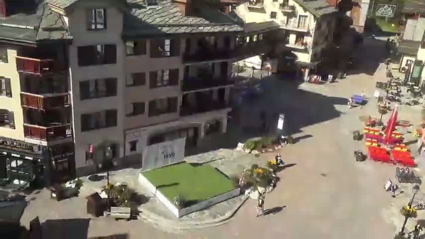 An aerial view shows a sunny town square with several buildings, a small grassy area, and some people walking around.