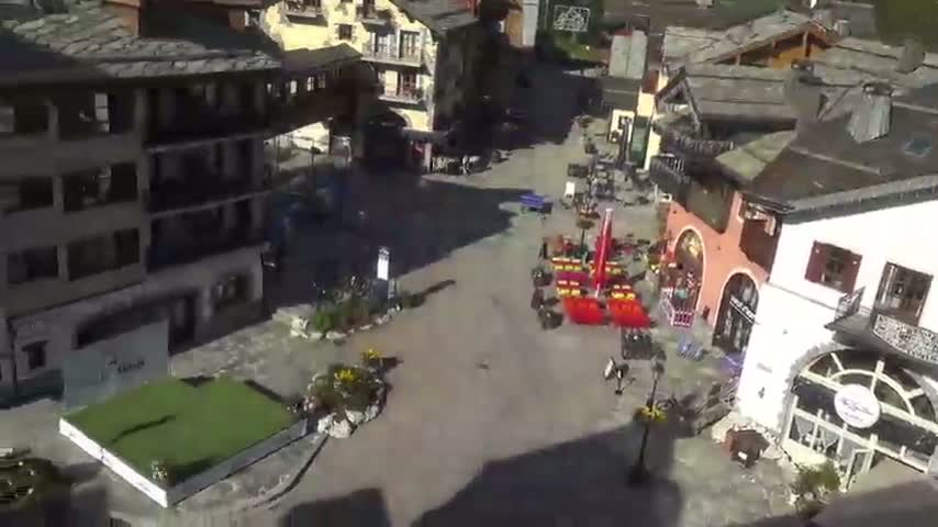 A sunny, high-angle view shows a town square with several buildings surrounding it, some outdoor seating, and a small patch of grass.