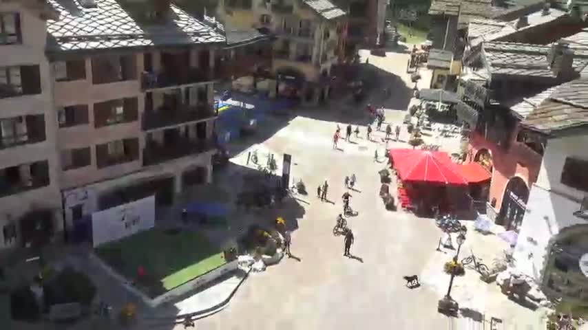 A sunny, high-angle view shows a town square with several buildings surrounding it, some outdoor seating, and a small patch of grass.