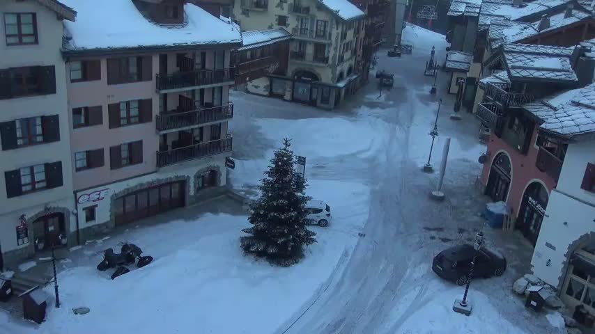 A snow-covered village square features a prominent decorated Christmas tree, surrounded by multi-story buildings with balconies and a few parked vehicles on the snowy streets, all under overcast and snowy conditions.
