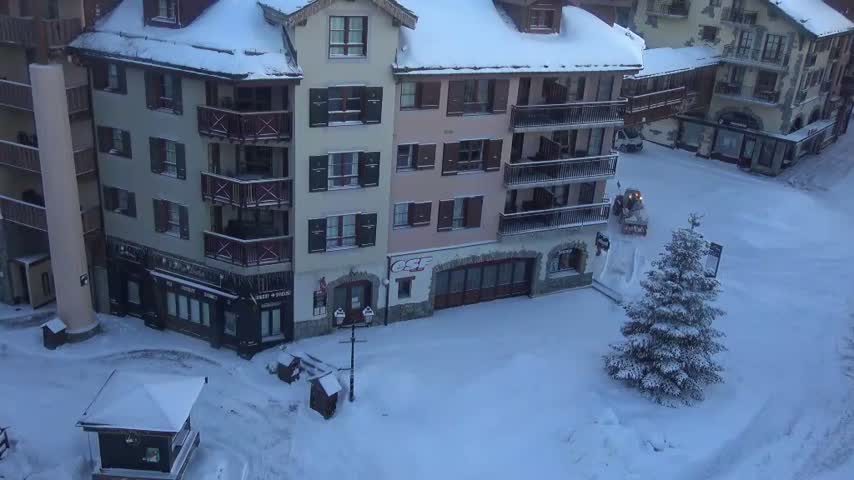An outdoor scene depicts a snow-covered village street flanked by multi-story alpine-style buildings with balconies, where a snowplow clears a path near a prominent evergreen tree, under cold and snowy conditions.