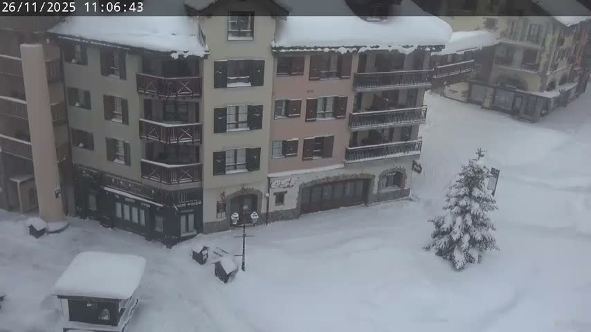 The image shows a snow-covered village square surrounded by multi-story buildings with balconies and ground-floor businesses, featuring a prominent snow-laden evergreen tree and completely blanketed ground under overcast, snowy conditions.