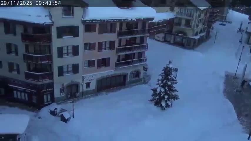 An elevated view reveals a peaceful village square deeply covered in fresh snow, with multi-story buildings and a prominent evergreen tree blanketed in white under cold, overcast weather.