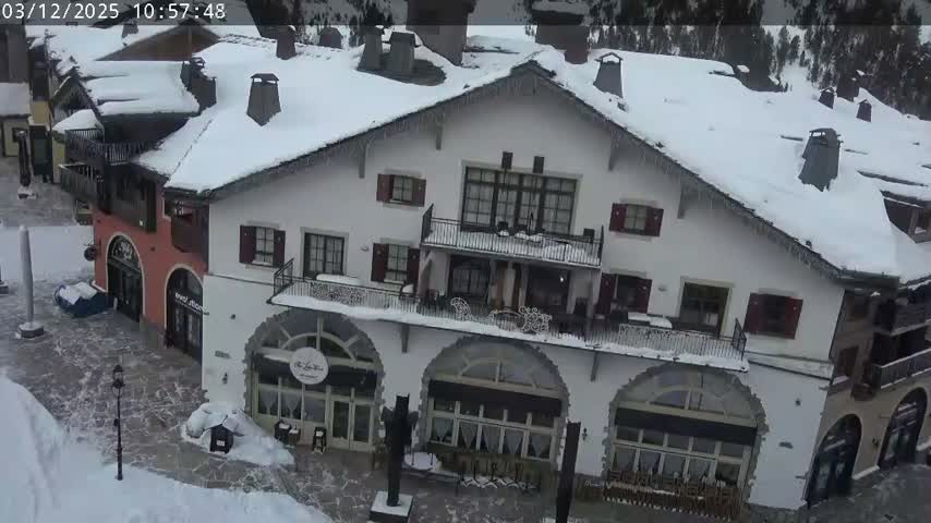 A charming European-style mountain resort village is depicted, blanketed in fresh snow with icicles hanging from roofs and partially cleared pathways, indicating cold, overcast winter weather.