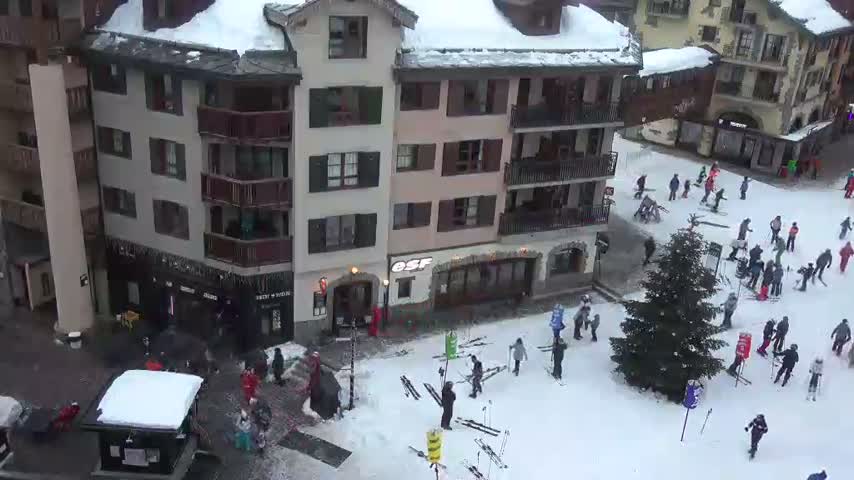 A bustling ski resort plaza is covered in snow on a clear winter day, with numerous people skiing and walking amidst multistory buildings and a large evergreen tree.