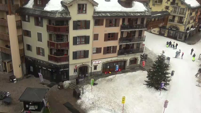 A bustling ski resort plaza is covered in snow on a clear winter day, with numerous people skiing and walking amidst multistory buildings and a large evergreen tree.