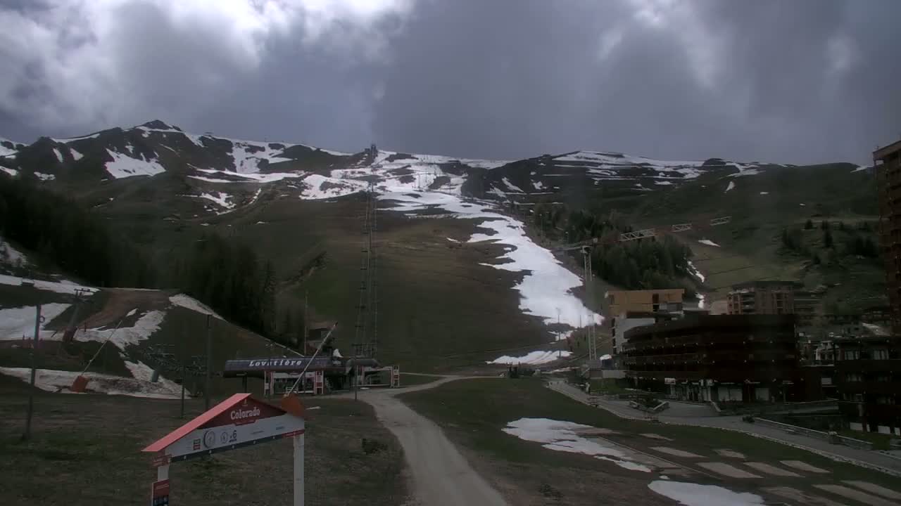 A mostly cloudy day reveals a mountainside ski resort with patchy snow, ski lifts, and buildings.