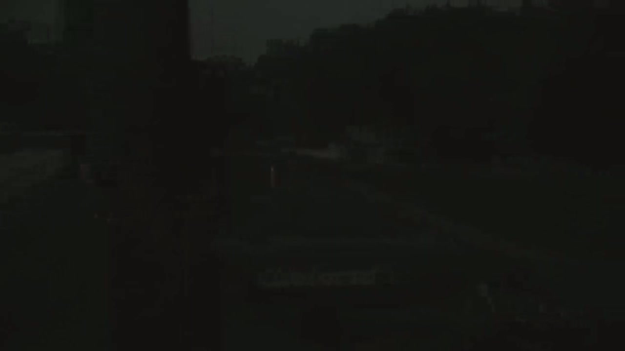A dark, nighttime scene shows a mostly obscured roadway with indistinct buildings and trees in the background under a dark sky.