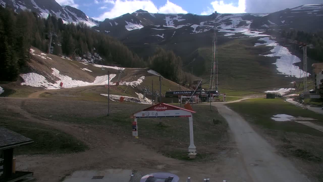 A partly sunny day at a ski resort shows mostly brown grassy hills with patches of snow, ski lifts, and mountains in the background.